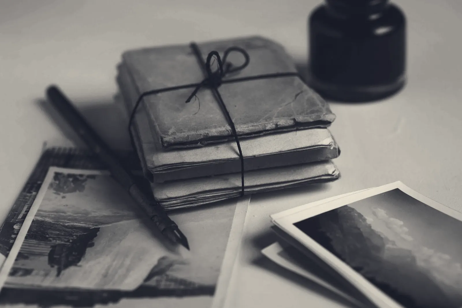 A stack of old letters tied with string, an ink bottle, a pen and a few vintage photographs on a table.