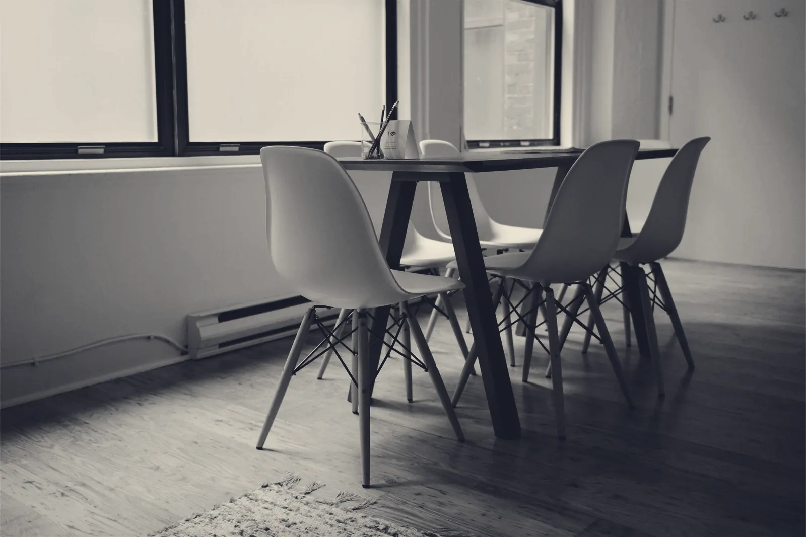 A quiet room with a wooden dining table and four chairs beside large windows, soft daylight falling on the floorboards.