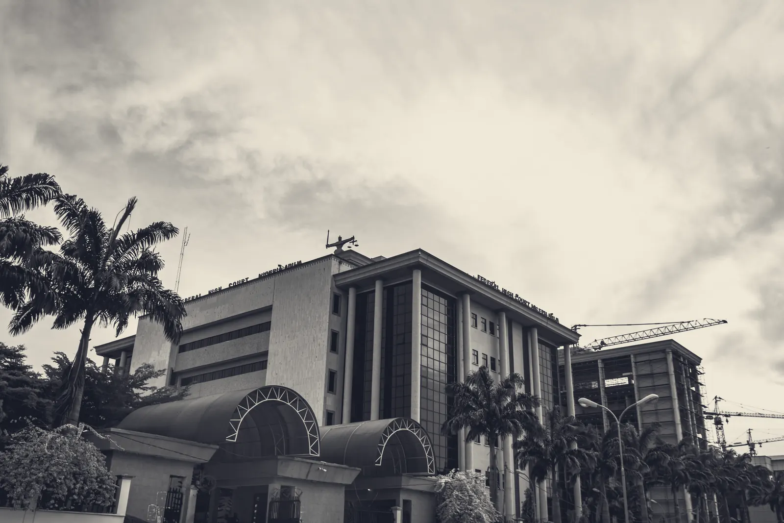 The Federal High Court complex in Abuja: a columned, multi-storey court building with arched entrance canopies, framed by palm trees under a cloudy sky.