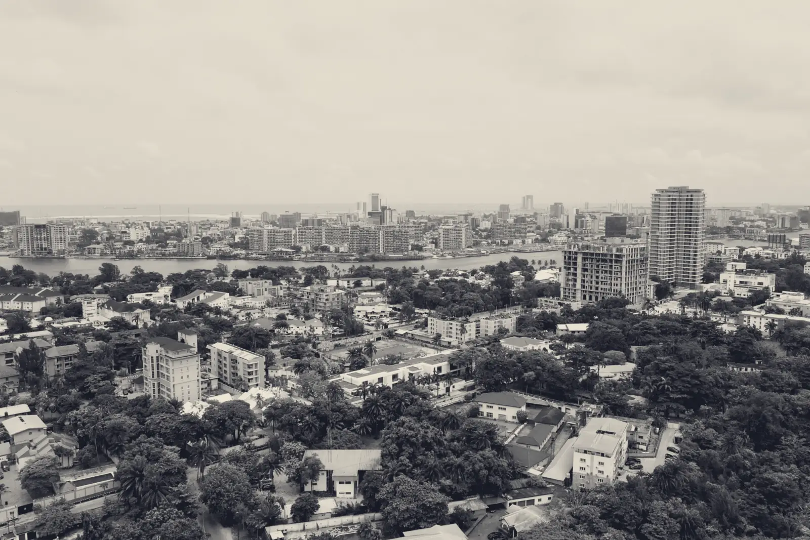 Aerial view over Ikoyi, Lagos: tree-lined residential streets and low-rise homes, new apartment towers under construction, and the city skyline across the water.