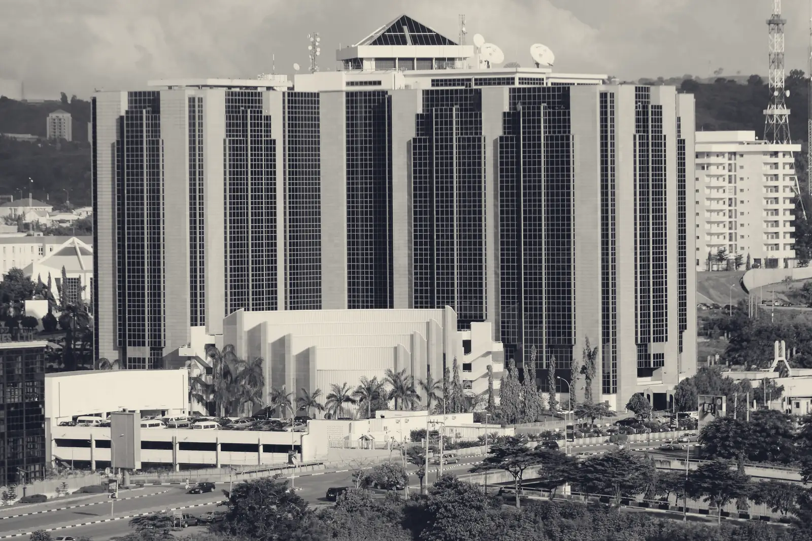 The Central Bank of Nigeria headquarters in Abuja, a large stepped tower of stone and dark glass, with palm trees and a road in front.