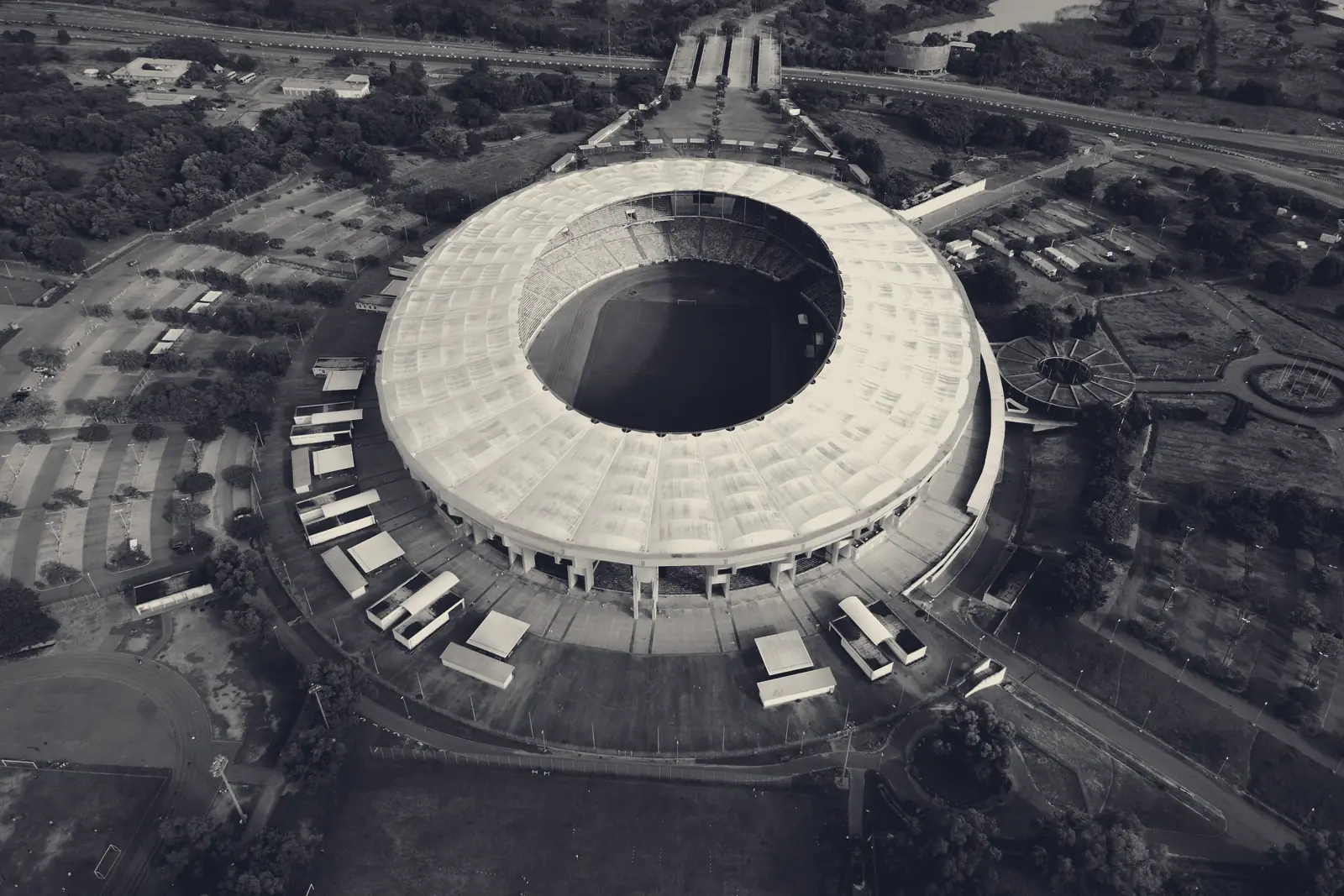 Aerial view of the Moshood Abiola National Stadium in Abuja: an oval bowl with a white roof ring around the pitch, surrounded by car parks and trees.