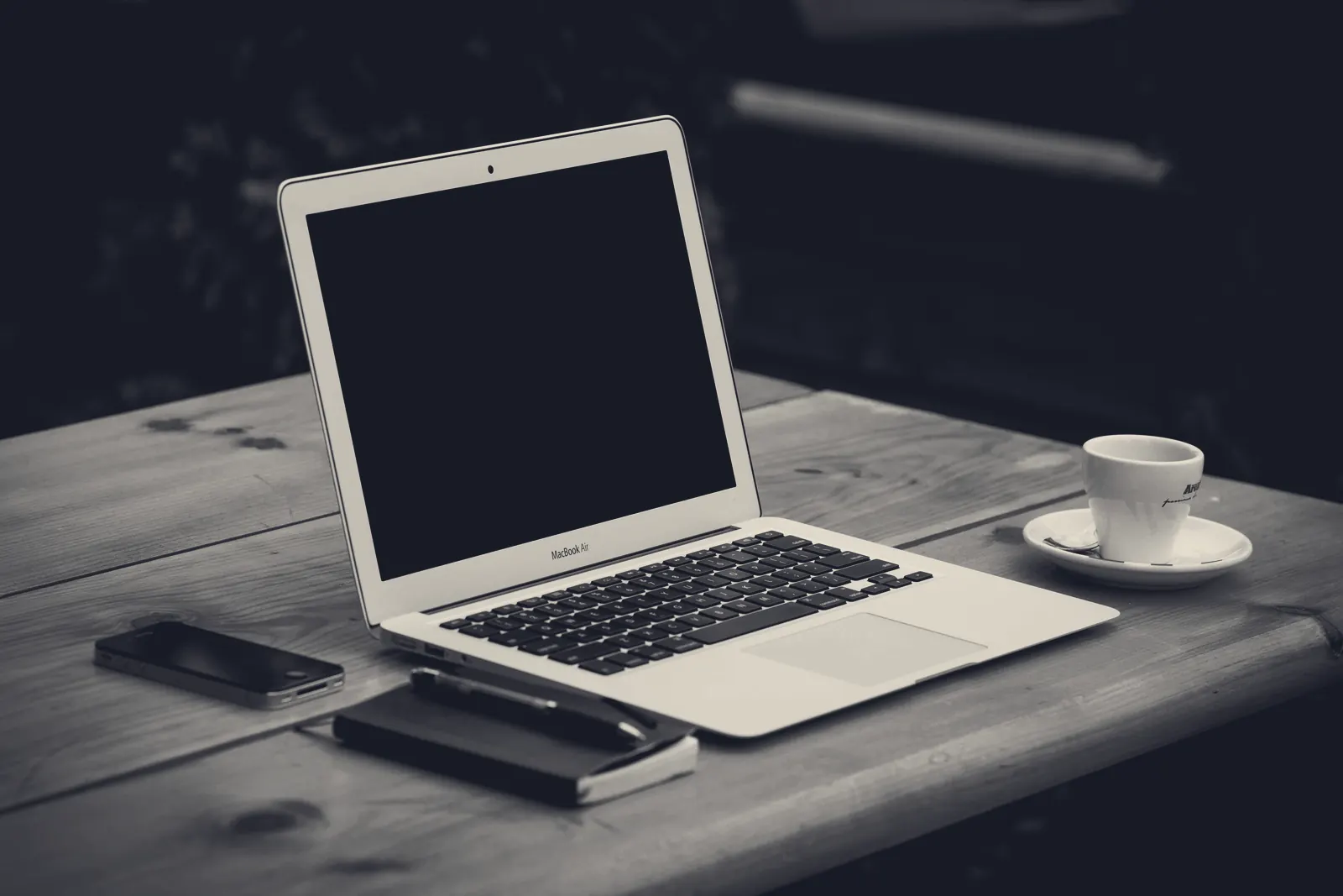 An open laptop on a wooden table with a notebook and pen, a phone and a cup of coffee, in a dim room.